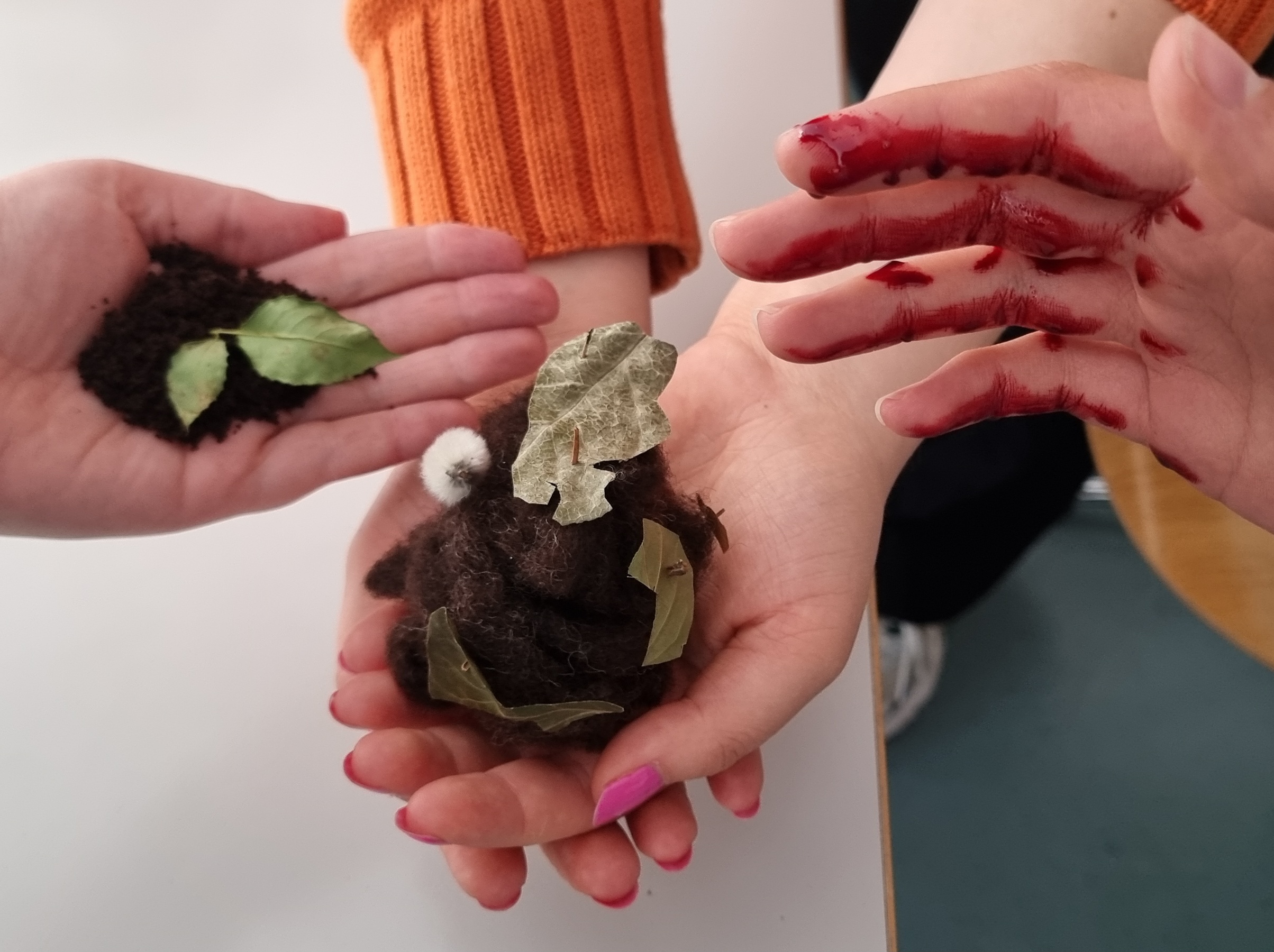 Foto von drei Händen, die Blätter und Wolle halten. Eine Hand ist mit Blut beschmiert. Anna Hielscher Harmlose Kunst