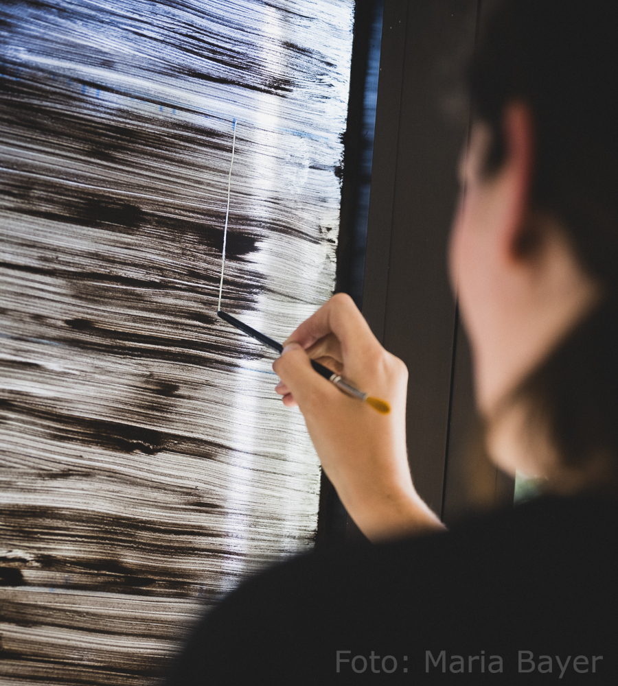 Foto von der Künstlerin Anna Hielscher, wie sie gerade in die Erdbeschichtung auf einer Glasscheibe kratzt. Harmlose Kunst Anna Hielscher
