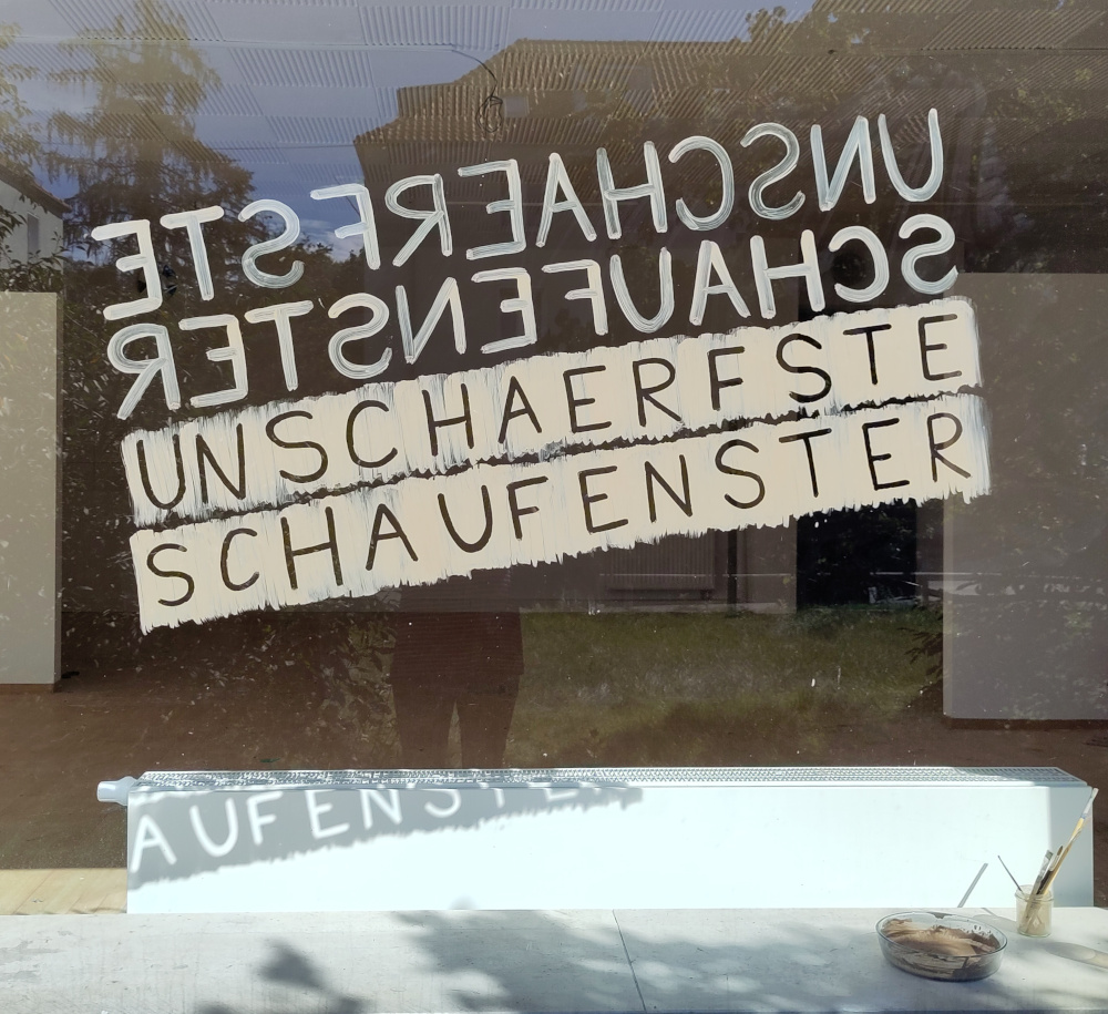 Foto von einer großen Fensterscheibe die mit Erde bemalt ist mit den Buchstaben SCHAUFENSTER UNSCHAERFSTE. Anna Hielscher Harmlose Kunst